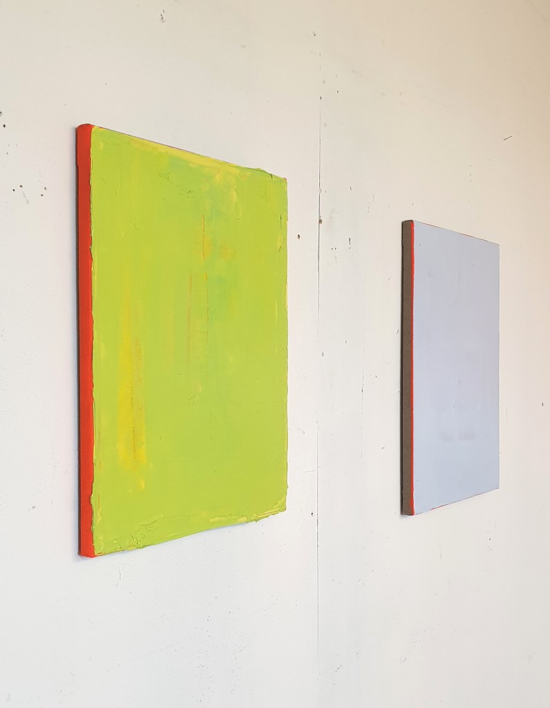 Two abstract paintings on a white wall, one dominated by a bright green color with a red border, and the other featuring a soft blue shade with a red edge.