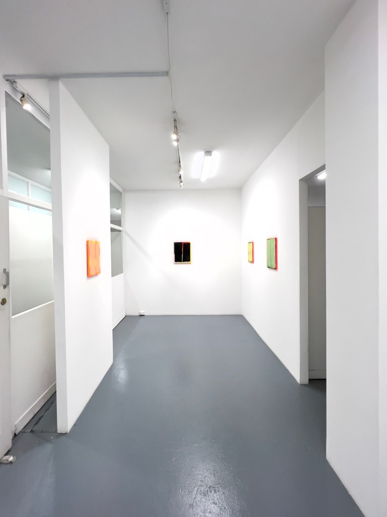 An art gallery interior featuring white walls and a grey floor, with abstract artwork displayed on the walls. The space is well-lit with track lighting.